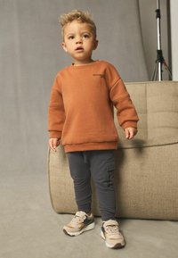 Sweat-shirt orange avec des poignets côtelés, pantalon cargo gris et baskets beiges. Tissu lisse, design ajusté et poches latérales sur le pantalon.