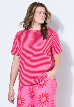 Frau mit welligem Haar, die ein rosa und rot gestreiftes T-Shirt und rosa Hosen mit roten Sonnenmustern trägt, schaut zur Seite mit einer Hand in der Tasche.