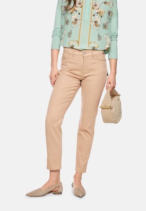 Woman wearing beige slim-fit pants, mint green floral blouse, beige pointed flats, and holding a small beige handbag by her side.
