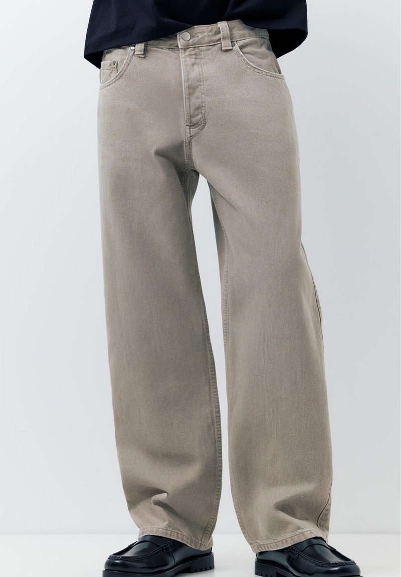 Jean beige à jambes larges porté avec des mocassins noirs, associé à un haut noir rentré, présenté sur un fond clair uni.