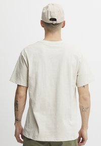 Cremefarvet bomulds t-shirt med afslappet pasform, korte ærmer og tagløs halsudskæring. Båret med en beige kasket og synlige arm-tatoveringer.