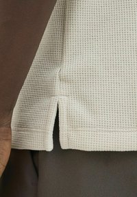 Tejido de punto tipo waffle en color beige claro con aberturas laterales, combinado con un material marrón más oscuro debajo. Presenta una superficie texturizada y colores contrastantes.