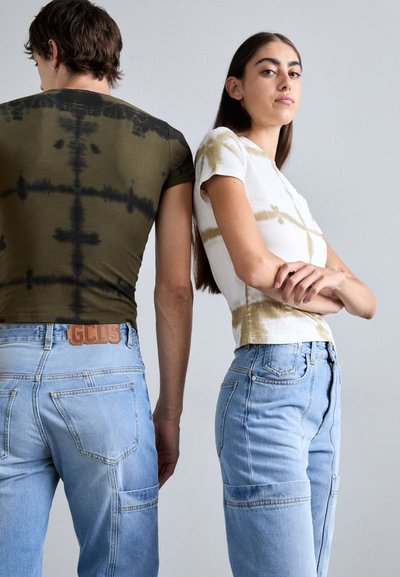 To personer iført tie-dye t-skjorter: en olivengrønn med mørkere mønstre, den andre hvit med lysebrune aksenter, kombinert med lyse denimjeans.