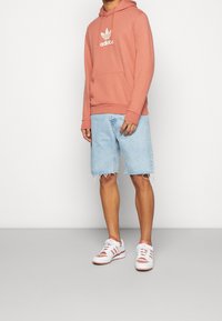 Ljusrosa hoodie med framficka, vit Adidas-logotyp. Ljusblå denimshorts med fransar. Vita sneakers med röda accenter och ränder.