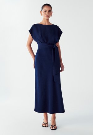 Navy blue, floor-length dress with short, wide sleeves, a defined waist tied with a belt, and a smooth texture. Thin strappy sandals.