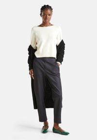 Maglione bianco lavorato a maglia, cardigan nero, pantaloni a righe fine e ballerine verdi. Design minimalista con texture e colori variati.