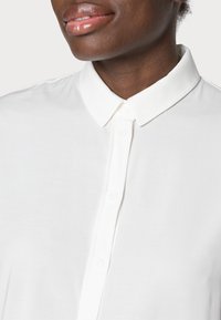 Chemise blanche à col, présentant une texture lisse, une fermeture à boutons et des lignes épurées. Gros plan sur le col et les détails des boutons supérieurs.