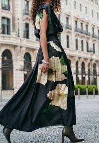 Frau, die in einem schwarzen Kleid mit großen weißen Blumenmustern und eleganten Stiefeletten auf einer Stadtstraße mit klassischer Architektur geht.