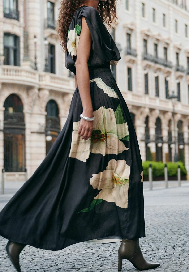 Frau, die in einem schwarzen Kleid mit großen weißen Blumenmustern und eleganten Stiefeletten auf einer Stadtstraße mit klassischer Architektur geht.