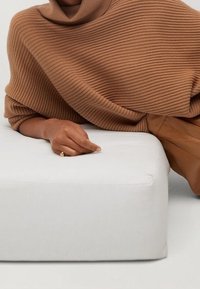 Pull en maille côtelée marron avec un col haut drapé sur un coussin beige. Une main portant une bague repose sur le bord du coussin.