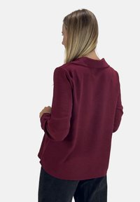 Bordeaux lange mouw blouse met een kraag en textuurstof, met een ronde zoom en een relaxte pasvorm. De achteraanblik toont schouderdetails.