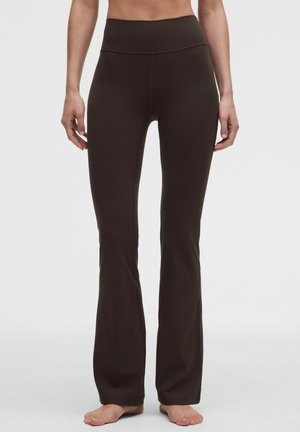 Mujer con pantalones acampanados ajustados de tiro alto en color marrón oscuro, de pie descalza frente a un fondo liso y claro.