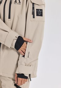 Beige wasserfeste Jacke mit dunklen Akzenten; mit Reißverschlüssen, Logo-Patches und einem verstellbaren Manschettendesign. Enthält eine Handtasche am Ärmel.