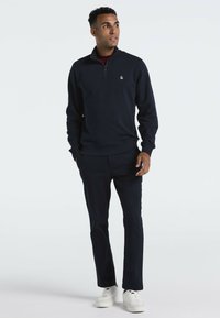 Marineblauwe pullover met een kwartrits en ribbels aan de mouwen, gecombineerd met bijpassende broek en witte sneakers. De stof lijkt glad en zacht.