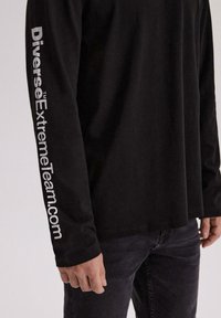 Czarny T-shirt z długim rękawem wykonany z bawełny, z białym napisem "DiverseExtremeTeam.com" wzdłuż lewego rękawa. Gładka faktura, luźny krój.
