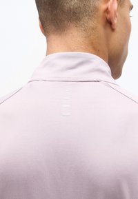 Η πλάτη και ο λαιμός άνδρα είναι ορατοί, φορεί ένα ανοιχτό μωβ πουκάμισο με ψηλό γιακά και πέντε μικρές λευκές οριζόντιες ραφές στο κέντρο.