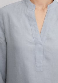 Blouse bleu clair avec un col mandarin, dotée d'une ouverture en v et d'une texture subtile, fabriquée à partir d'un tissu léger et respirant.