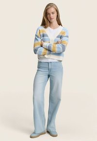 Kötött kardigán kék, sárga és fehér csíkokkal, V-nyakkivágással és gombos lezárással. Világoskék bővülő farmerral párosítva.