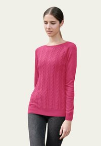 Pull en tricot torsadé rose à manches longues et col rond, fabriqué dans un matériau doux, associé à un jean skinny foncé.