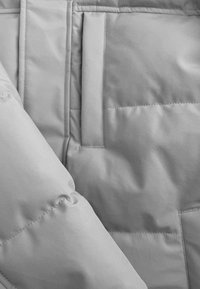 Lichtgrijs pufferjackstof met een gladde textuur, voorzien van gewatteerde voering, een patchzak en gestikte details op de mouw.