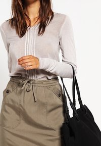 Top grigio chiaro a maniche lunghe con piastre verticali, abbinato a una gonna verde oliva a vita alta con tasche anteriori e cordoncino, insieme a una borsa a tracolla nera.