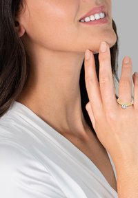 Zlatý zásnubný prsteň s centrálnym okrúhlym diamantom, na ruke dotýkajúcej sa brady. Modelka má dlhé vlasy a nosí biely top.
