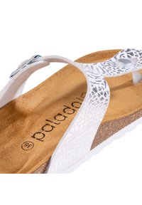 Sandalias blancas tipo thong con tira texturizada, acentos metálicos y plantilla de corcho. Presenta un patrón en relieve y plantilla con la marca.