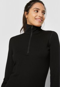 Czarny, długi rękaw, top z wysokim kołnierzem i pół zamkiem. Gładka tekstura, dopasowany kroj i minimalistyczny styl bez widocznych logo lub wzorów.