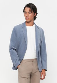 Man wearing light blue blazer over white shirt and beige pants, looking to the side with one hand in pocket against plain background.