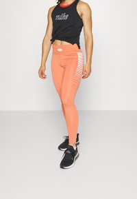Zwarte mouwloze top met het "nike" logo, gecombineerd met nauwsluitende, middelhoge koraalkleurige leggings met witte diagonale strepen aan één kant. Zwarte sportschoenen.