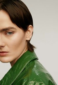 Chaqueta verde brillante con un cuello estructurado, llevada con pequeños pendientes: uno de piedra preciosa circular y una barra de metal elegante. Textura de piel suave.