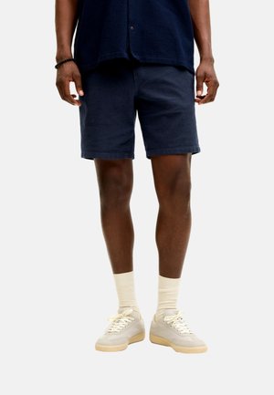 Hombre vistiendo pantalones cortos azul marino, camiseta azul marino, calcetines crema y zapatillas beige, de pie contra un fondo blanco.