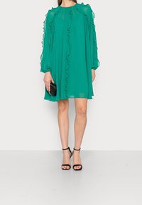 Zelené šifónové šaty s volány podél výstřihu a rukávů, střední délky, doplněné černými sandály na podpatku a černou kabelkou.
