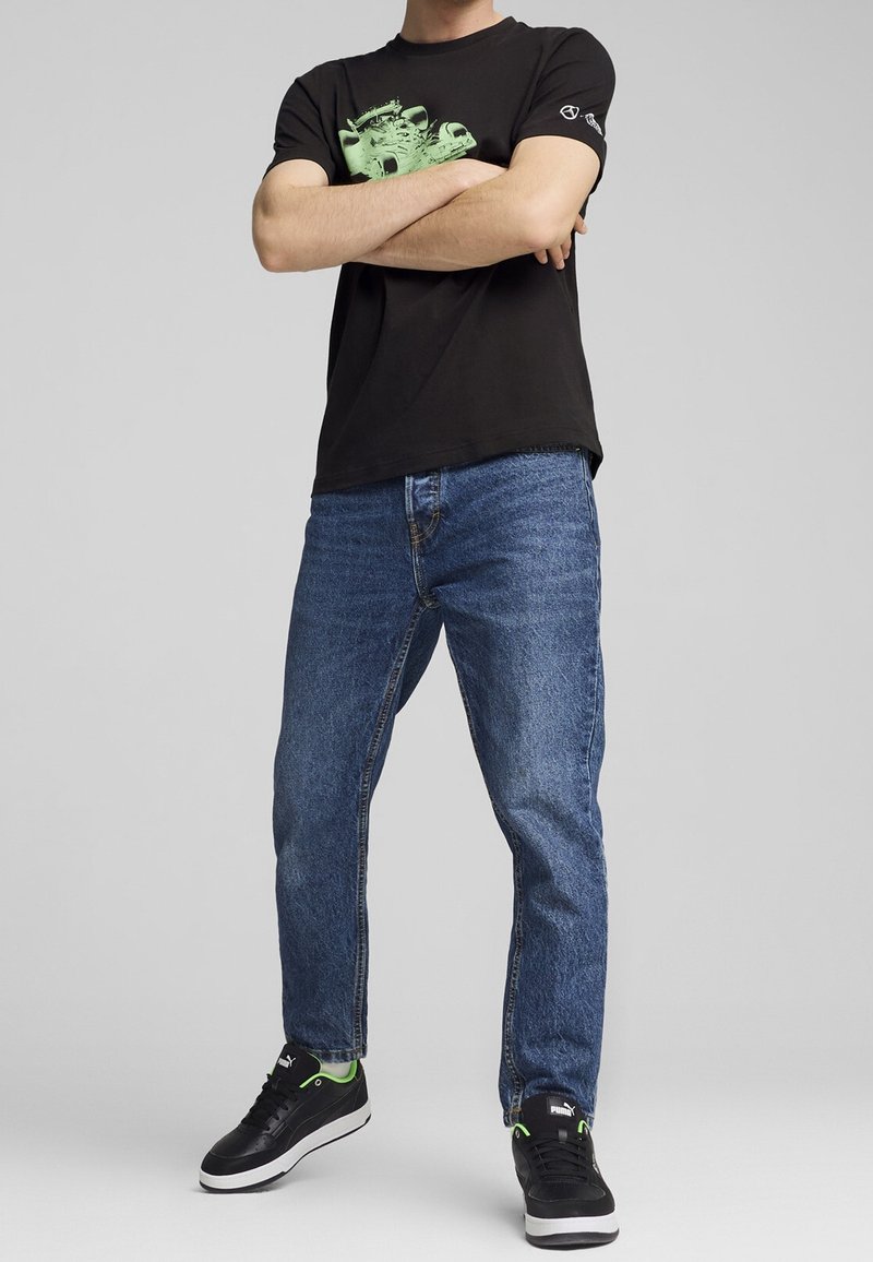 Personne portant un t-shirt graphique noir, un jean bleu et des baskets noires avec des accents verts, les bras croisés sur un fond gris.