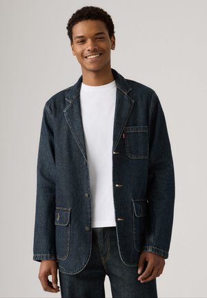 Jachetă din denim închis, cu două buzunare frontale, guler cu rever și nasturi. Pur tată peste un tricou alb, are cusături vizibile și o croială lejeră.