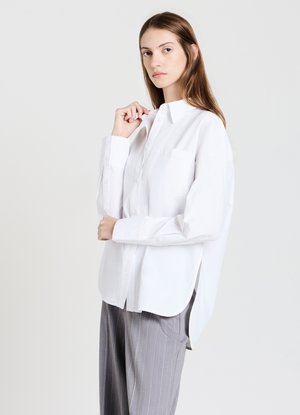 Donna che indossa una camicia button-up bianca oversize e pantaloni grigi a righe, che sistema il colletto della camicia mentre guarda in avanti su uno sfondo bianco.