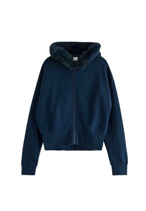 Ung kvinde i marineblå jakke med lynlås og pelskrave, blomstret top, matchende bukser og lyse beige sneakers går fremad.