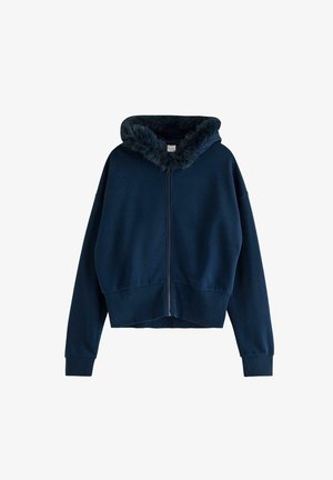 Ung kvinde i marineblå jakke med lynlås og pelskrave, blomstret top, matchende bukser og lyse beige sneakers går fremad.
