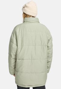 Die Person, die von hinten zu sehen ist, trägt eine hellgrüne Puffjacke und eine cremefarbene Strickmütze, unter der langes blondes Haar sichtbar ist.