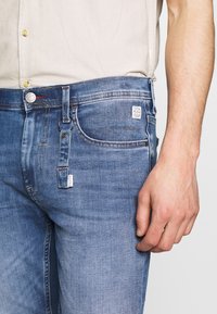 Blå denimjeans med smal passform, knappgylft, fem fickor och en logotyppatch bak. Har subtila whiskeringar och sömnadsdetaljer.