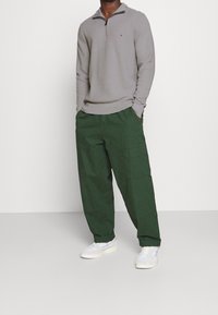 Maglione grigio lavorato a maglia con colletto chiuso da zip, abbinato a pantaloni cargo larghi di colore verde scuro e sneakers bianche, che mostrano un fit rilassato e uno stile casual.