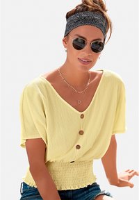 Blusa gialla strutturata con maniche corte, scollatura a V e tre bottoni in legno sul davanti. Vita arricciata e orlo a volant. Occhiali da sole tondi.