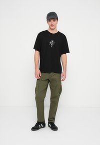 Schwarzes Kurzarm-T-Shirt mit kleinem weißem Blumenmotiv, kombiniert mit olivgrünen Cargo-Hosen. Schwarze Sneaker und eine graue Kappe vervollständigen den Look.