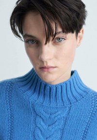Blauer Strickpullover mit hohem, geripptem Kragen, strukturierten Mustern und einem zentralen Zopfdesign. Weiche Textur und eng anliegender Stil.