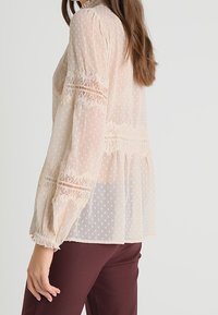 Transparente cremefarbene Bluse mit Punkten, Spitzenverzierungen und Puffärmeln. Enganliegende Passform mit einem gerafften Bund und elastischen Bündchen. Getragen mit burgunderfarbenen Hosen.