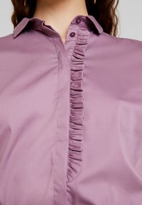 Primo piano di una persona che indossa una camicia malva con un bottone plissettato e colletto, che mostra la parte superiore del torace e il collo.