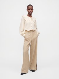 Femme portant un chemisier crème à manches longues avec un col à volants et un pantalon beige à larges jambes à carreaux, les mains dans les poches.