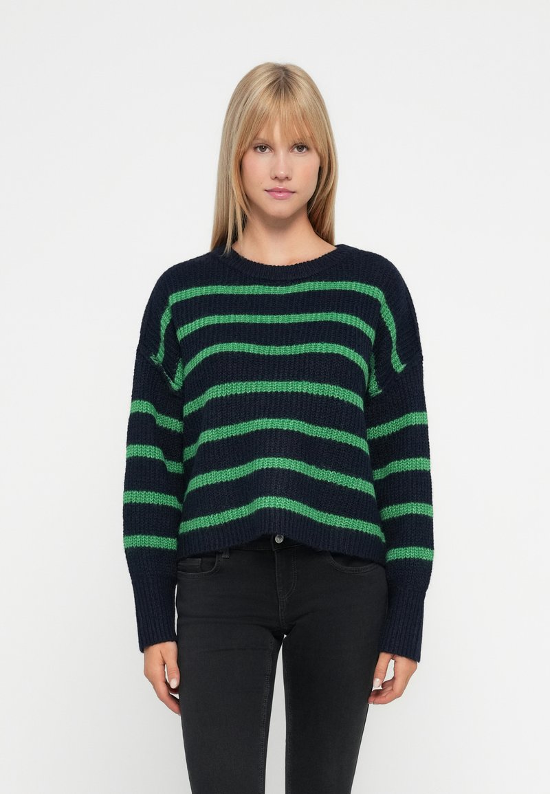 Joven mujer vestida con un suéter azul marino con rayas verdes horizontales y jeans negros, de pie frente a un fondo blanco liso.