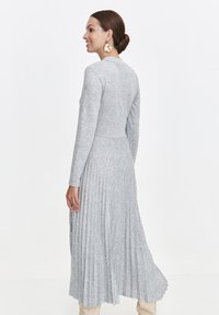 Frau mit einem Dutt, die große goldene Blütenohrringe und ein langes, graues, plissiertes Kleid trägt, lächelt und schaut nach links.