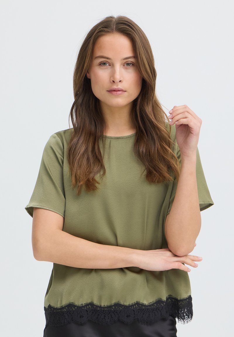 Femme aux longs cheveux bruns portant un haut à manches courtes vert olive avec une bordure en dentelle noire, debout avec une main levée près du visage.